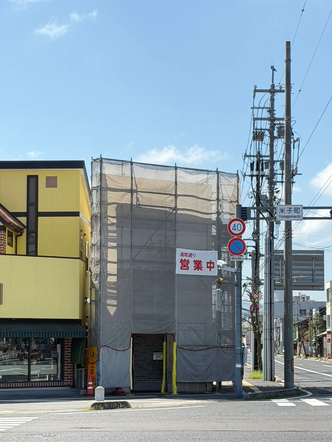 外壁工事のため足場と養生シートを設置したIEKAKE店舗の外観。営業中の看板が出ている
